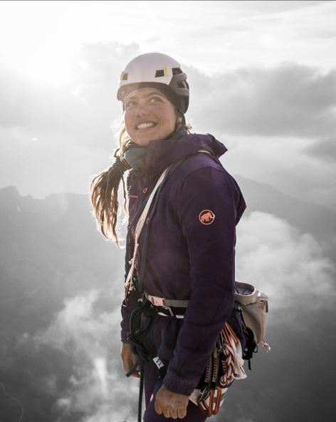 A climber in a helmet and Mammut gear stands on a mountain, smiling, with a cloudy sky and the sun shining behind her.