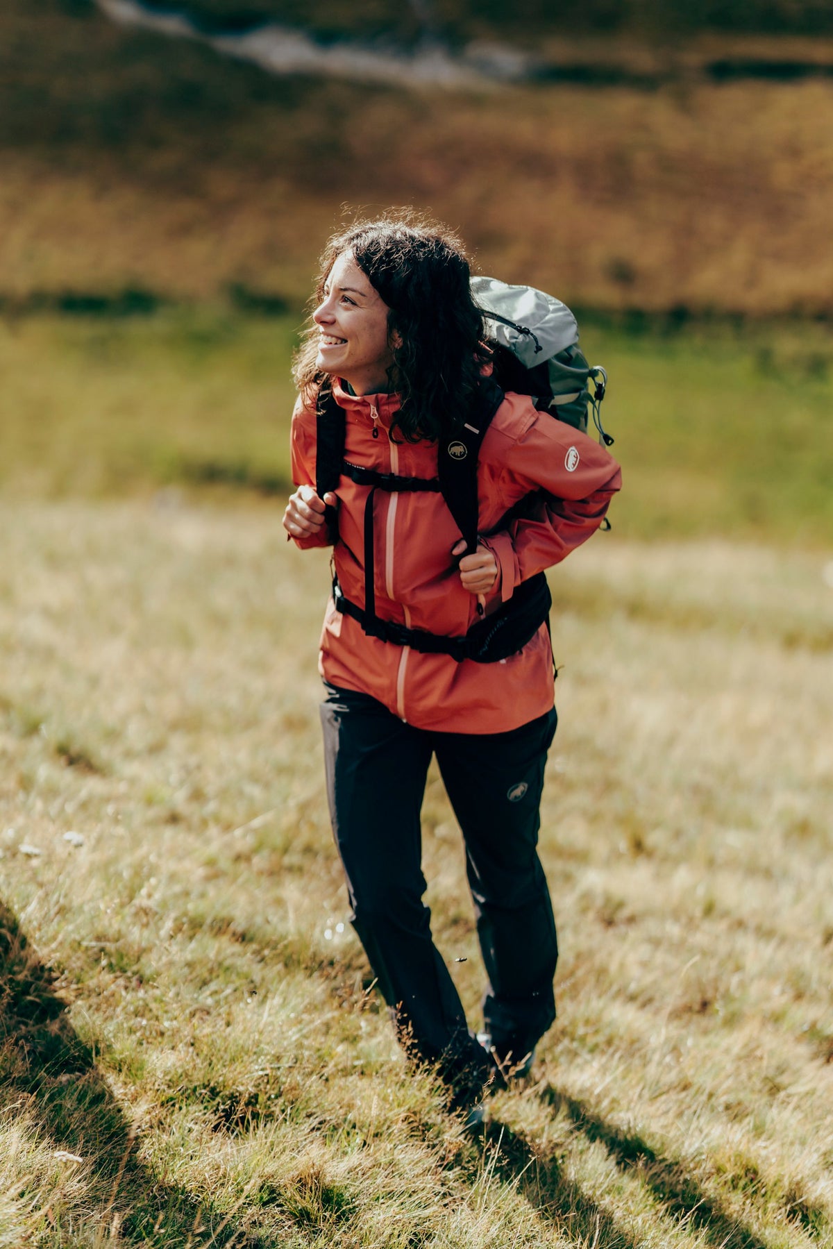 Mammut-clad hiker in a red jacket and green Mammut backpack enjoying a trek on a grassy hill.