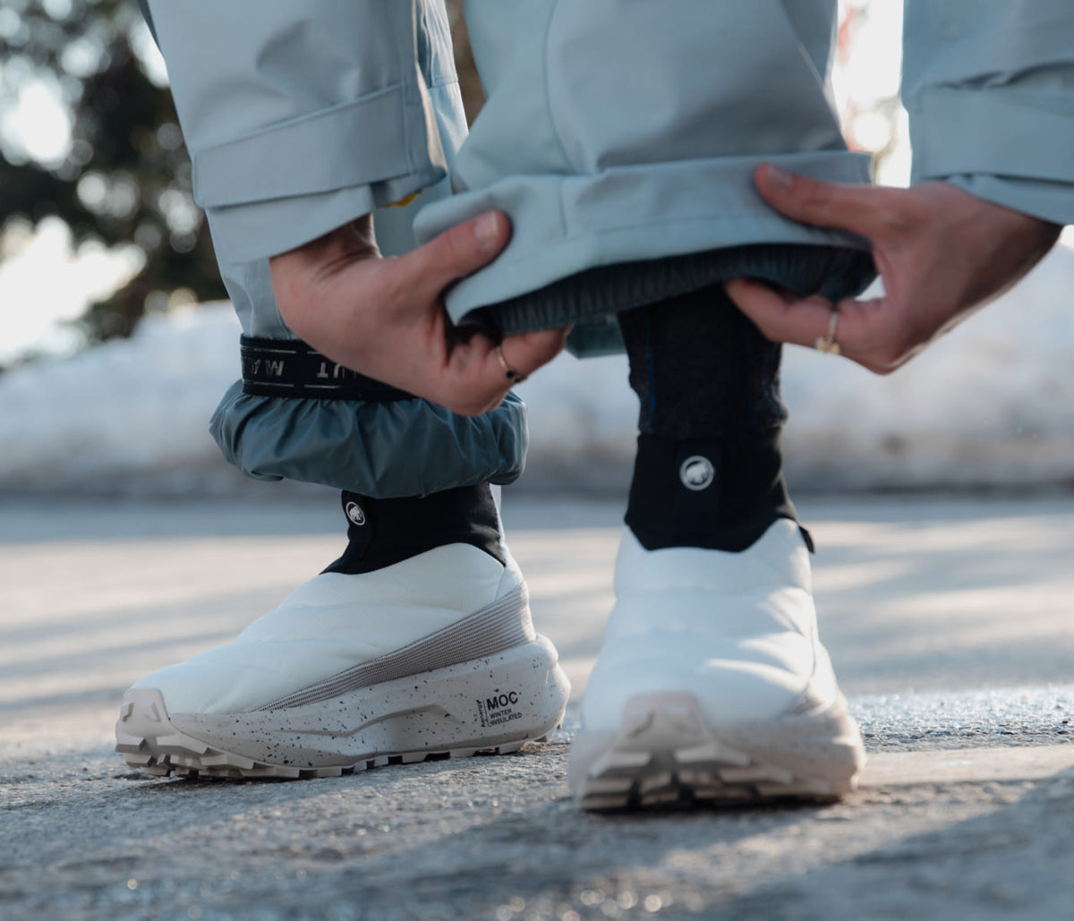 Close-up of a person adjusting pant leg over white Mammut outdoor shoes with black gaiters, standing on pavement.