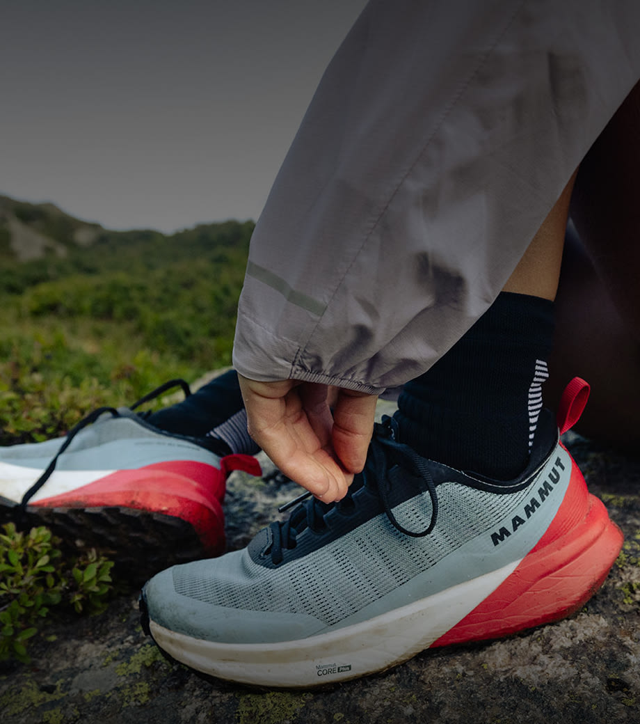 Close-up of a person tying laces on a gray and red Mammut trail running shoe outdoors on a rugged, rocky mountain path.
