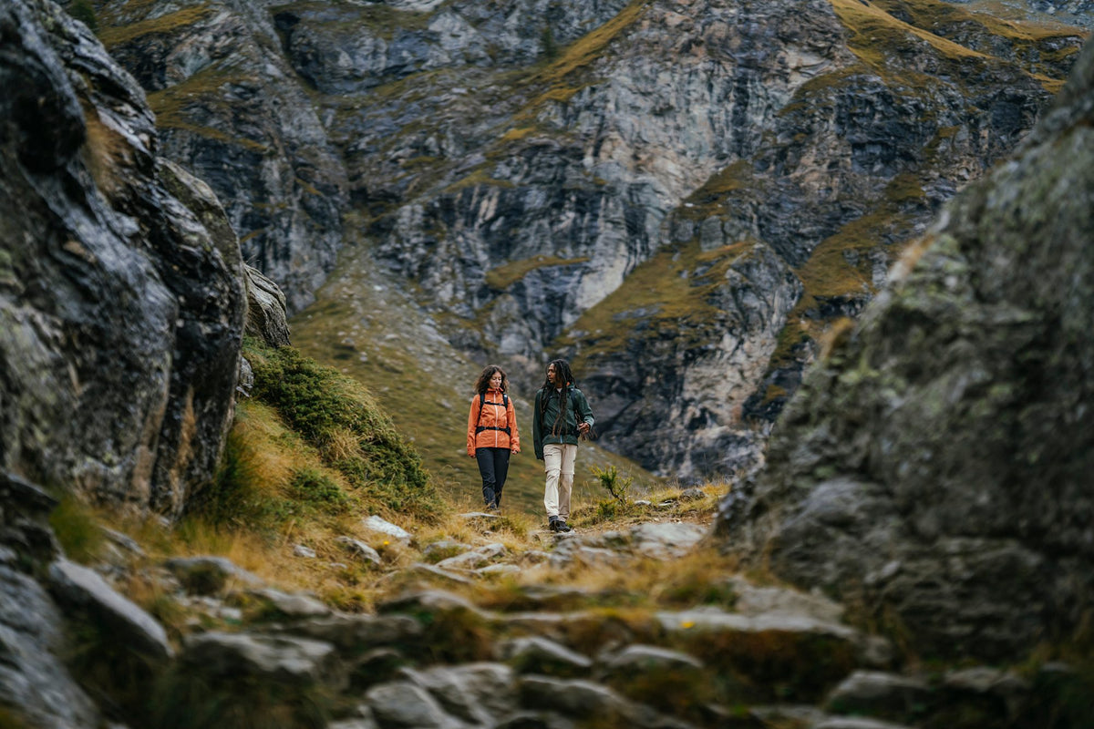 Two hikers wearing Mammut gear navigate a rocky mountain trail with steep cliffs and rugged terrain in the background.