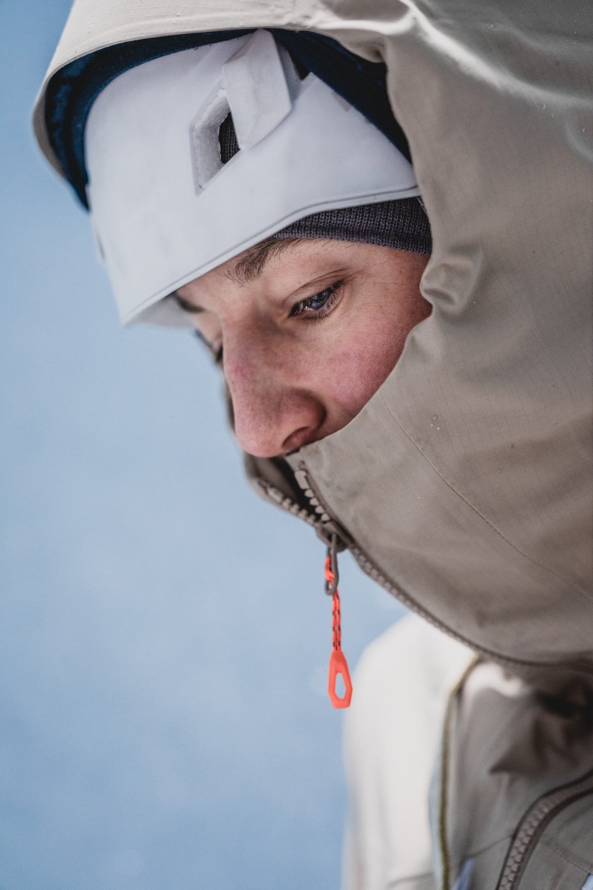 Individual wearing a beige Mammut hooded jacket with face partially obscured, gazing downward against a vibrant blue backdrop.