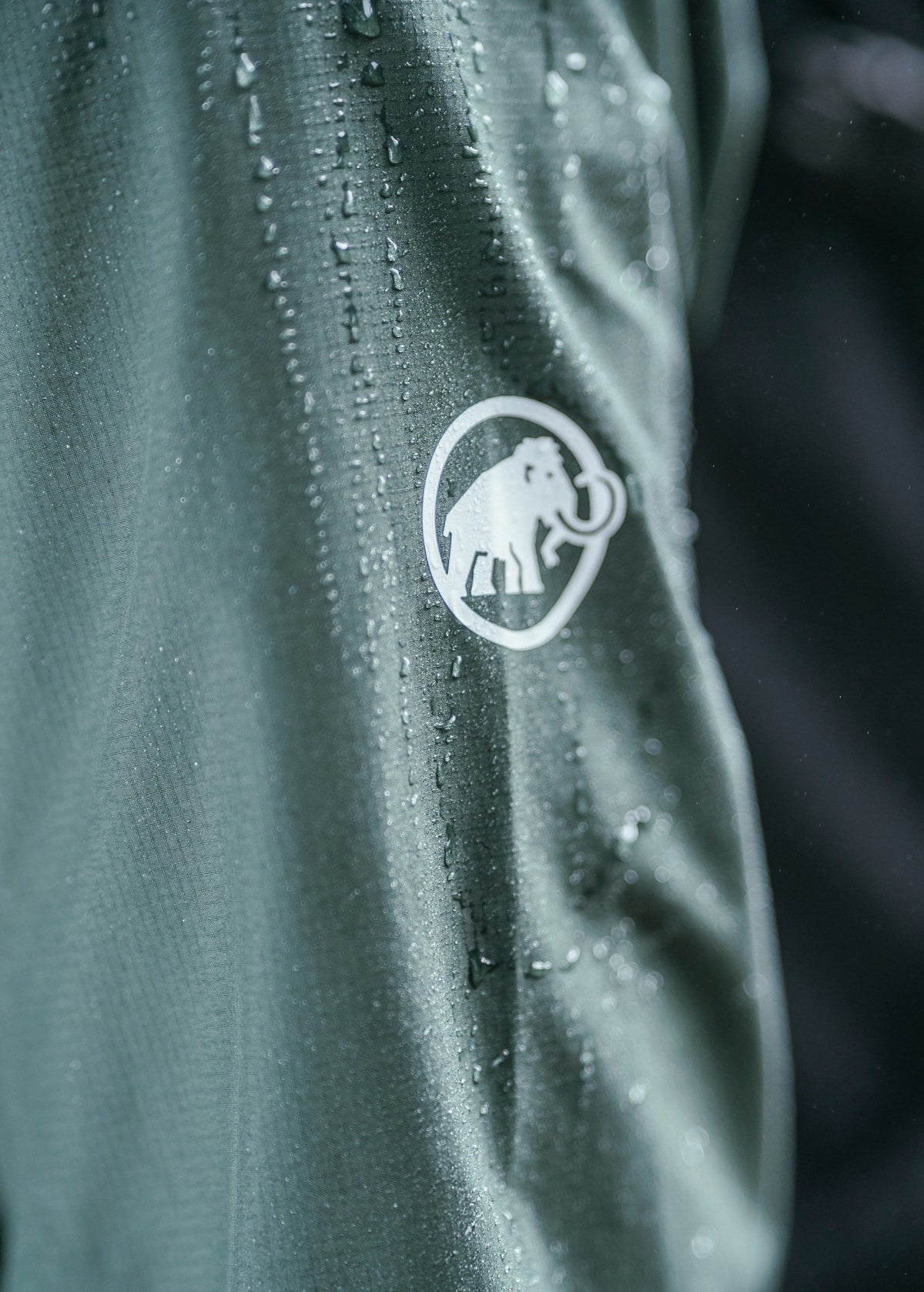 Close-up of dark green Mammut waterproof fabric with water droplets and a white woolly mammoth logo.