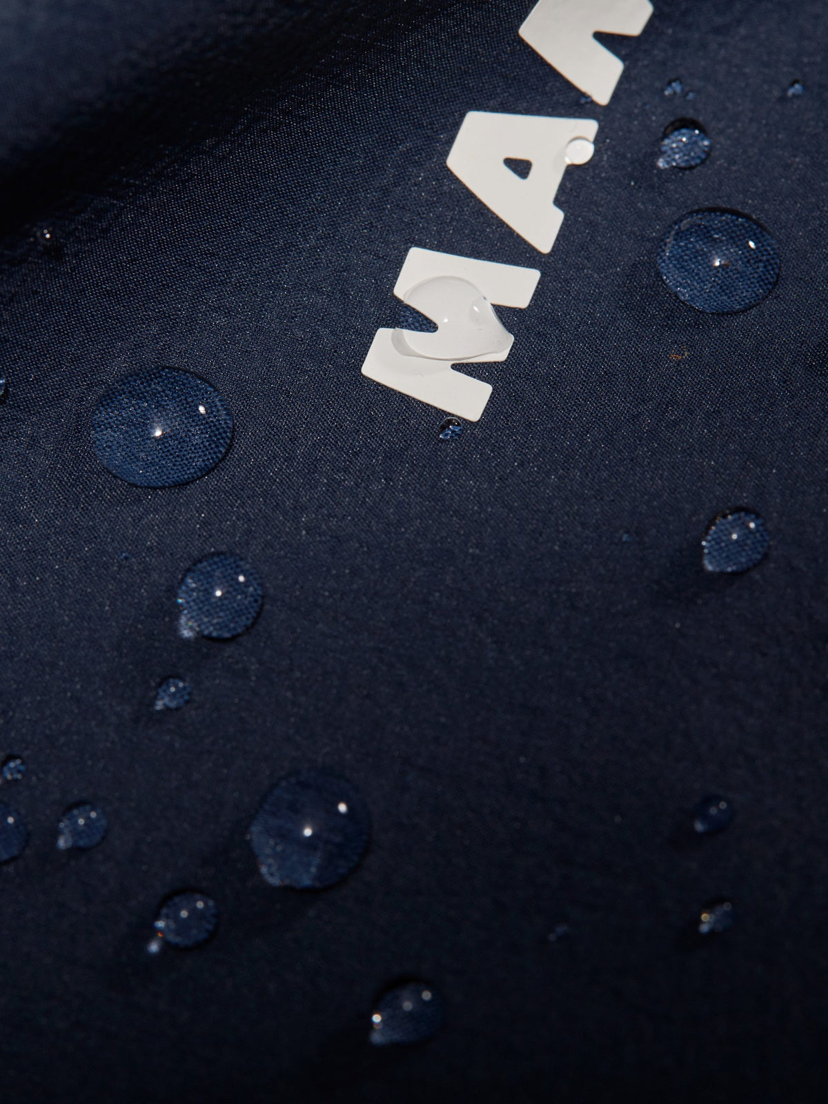 Close-up of water droplets on a dark blue Mammut jacket fabric, featuring part of the white Mammut logo.