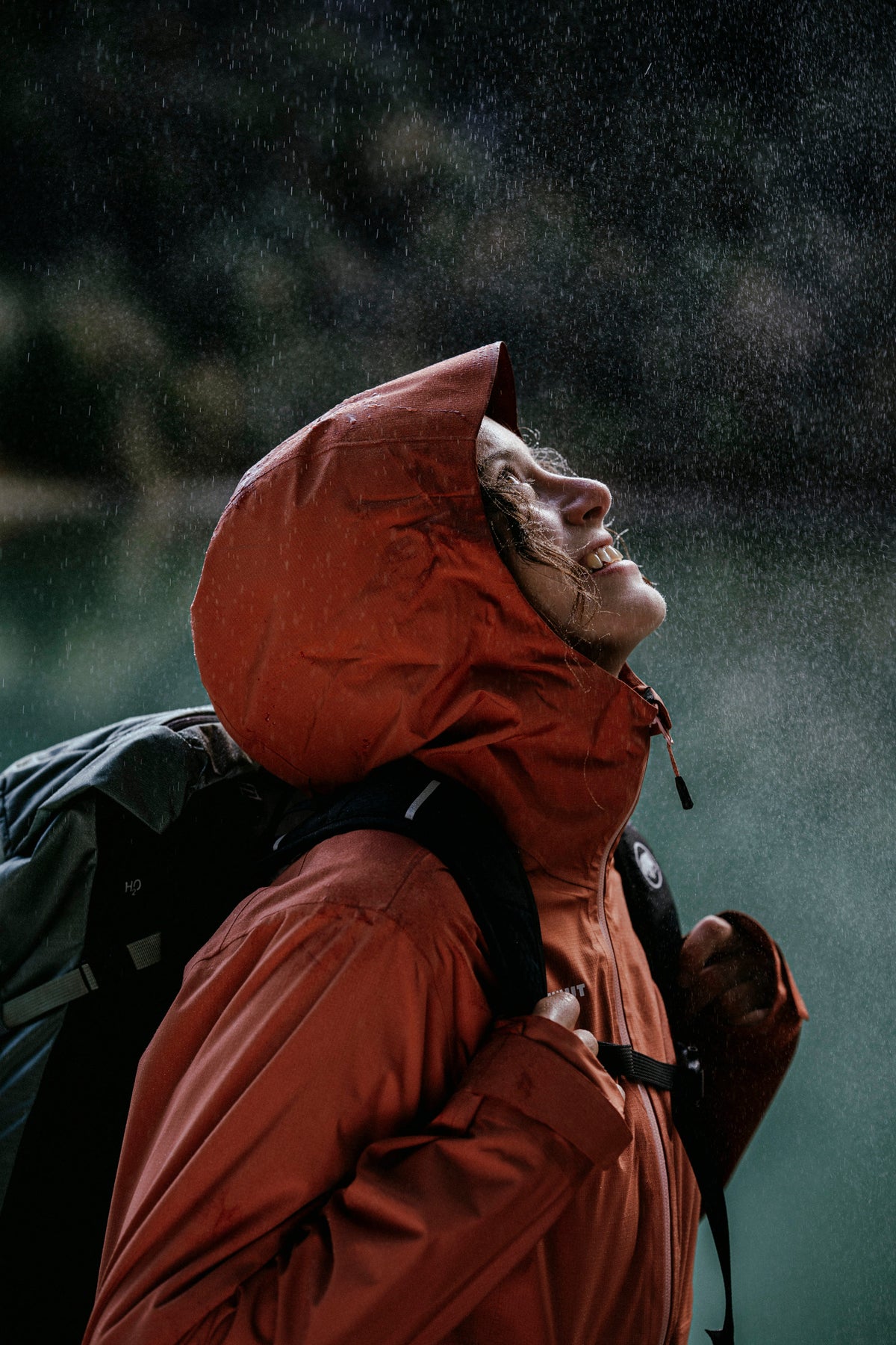 Person in a Mammut orange rain jacket and hood, smiling and looking up at the sky, wearing a backpack, in the rain.