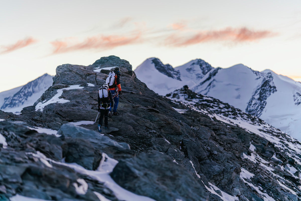 Two climbers with Mammut backpacks traverse a rocky mountain ridge, surrounded by snow-covered peaks at dawn.