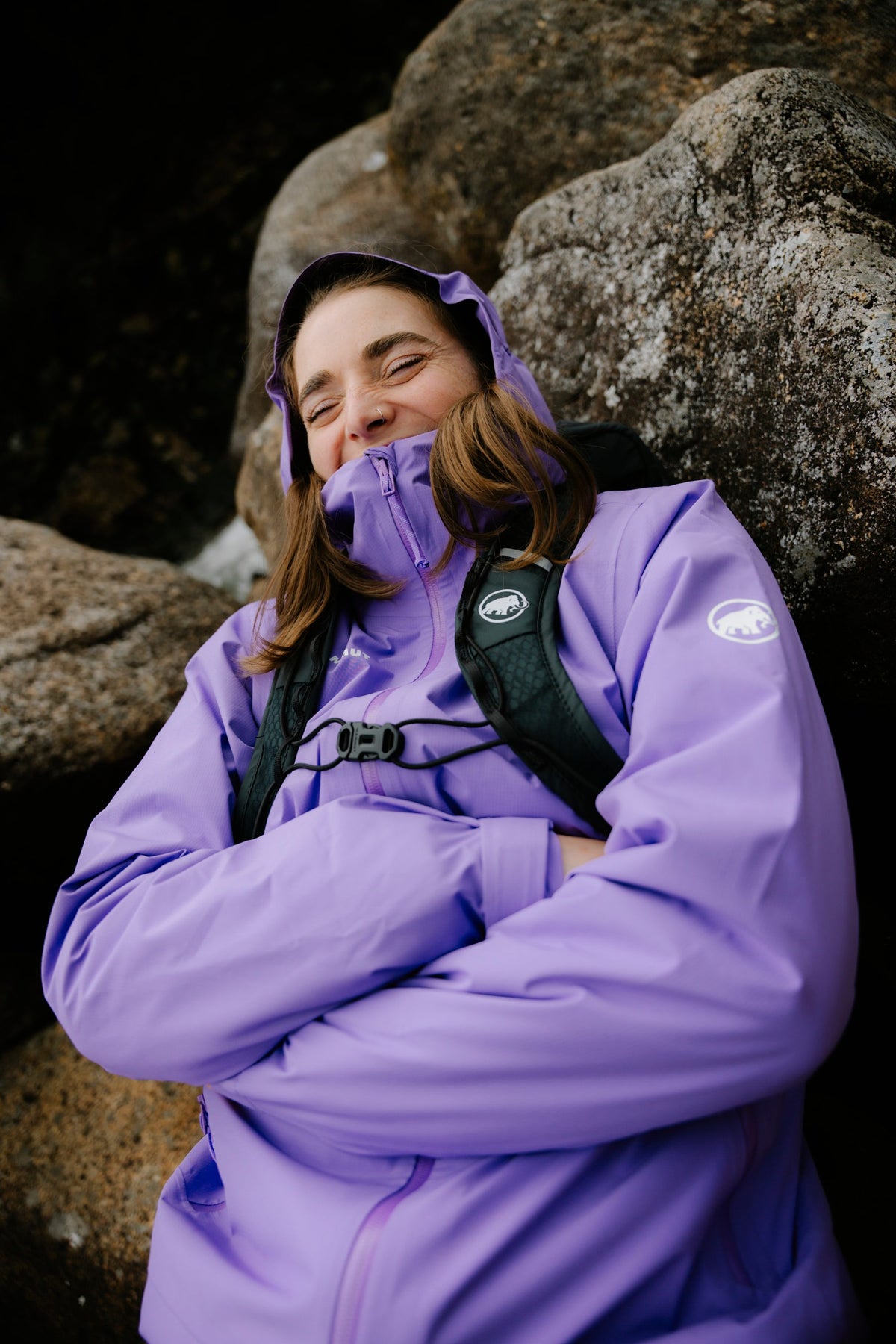 A person in a Mammut purple jacket smiles with arms crossed, leaning against large boulders in an outdoor mountain setting.
